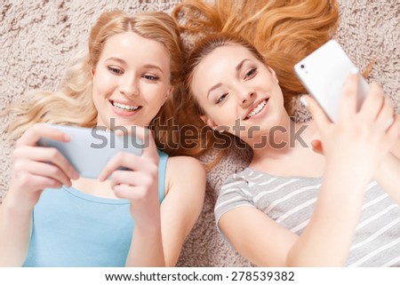 Selfie time. Two young beautiful blond girls wearing pajamas lying on the light floor, smiling and making selfie top view