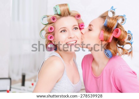 Friendship. Young blond girl kissing her best friend at her cheek smiling and wearing pajamas and colorful hair rollers at home party in the light room