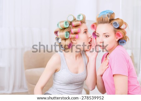 Did you know. Young blond girl sharing a secret to her best friend looking very surprised wearing pajamas and colorful hair rollers at home party in the light room