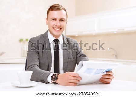 Read with smile. Smiling businessman holding newspaper in white decorated in kitchen.