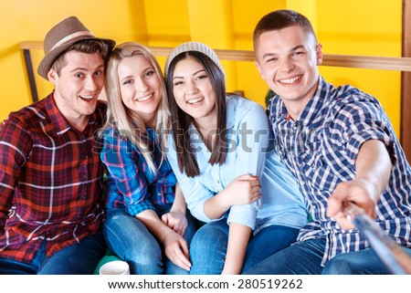 Young carefree happy students wearing casual clothes making photos with a selfie stick and smiling in a yellow room