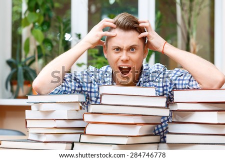 Forgot something important. Portrait of a handsome smart student sitting behind a big stack of books in a library, holding his head opening his mouth and looking very shocked