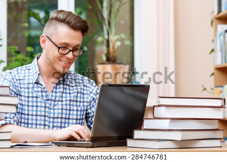 Portrait of a handsome smart student wearing glasses and blue checkered shirt sitting at the table in a library and working on his computer smiling, a stack of books and a laptop on the table