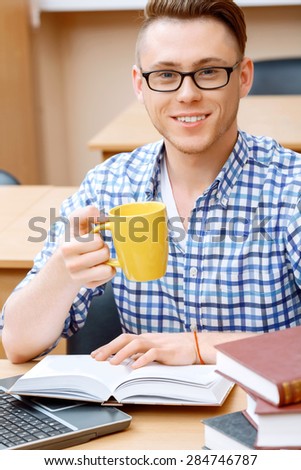 Portrait of a handsome smart student wearing glasses sitting at the table in a library and working on his computer and drinking tea from yellow cup smiling, a stack of books on the table