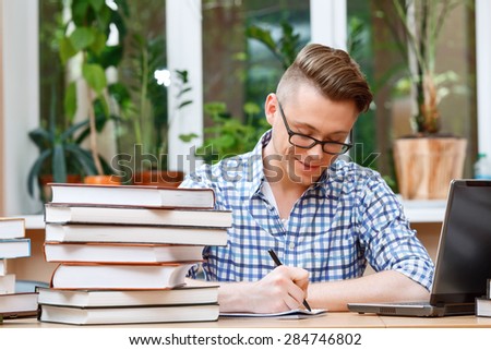 Portrait of a handsome smart student wearing glasses and blue checkered shirt sitting at the table in a library and making some notes in his notebook, a stack of books and a laptop on the table