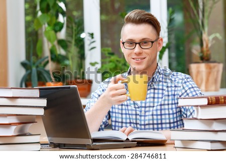 Portrait of a handsome smart student wearing glasses sitting at the table in a library and working on his computer and drinking tea from yellow cup smiling, a stack of books on the table