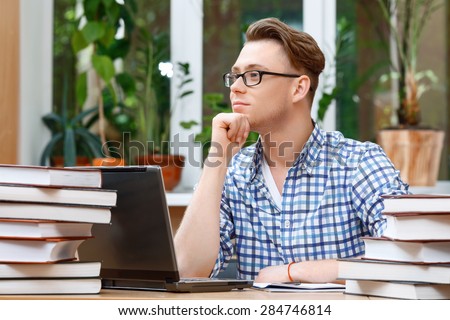 Portrait of a handsome smart student wearing glasses sitting at the table in a library and holding his hand on his chin thinking of something, a stack of books and a laptop on the table