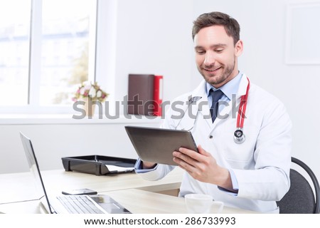 Feeling healthy.  Smiling cardiologist holding the laptop and looking at it  while  sitting in consulting room.