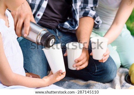 Close up photo of hands of mother and daughter holding white paper cups waiting for father pouring tea from thermos, sitting on a plaid on a green grass, selective focus