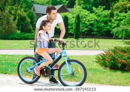 Portrait of a happy caring father teaching his small pretty daughter riding a bicycle in a green park ,smiling full length