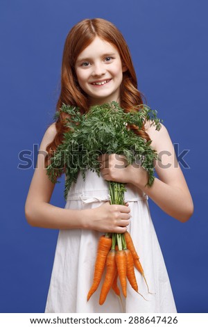 Feeling healthy. Pretty red-haired girl holding a bunch of  carrot in two hands and smiling while standing isolated on blue background.