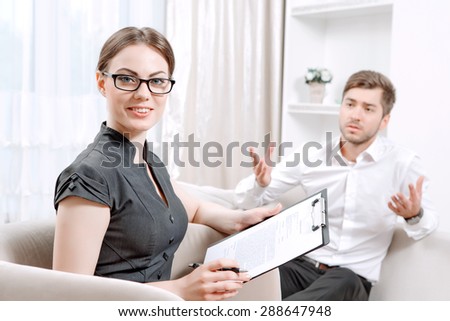 Young man wearing a white shirt sitting on a couch telling his problems and gesticulating, psychologist with clipboard listening to him and looking at us during therapy session, selective focus