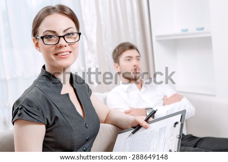 Portrait of a beautiful woman psychologist wearing glasses and black blouse sitting at the light doctor office smiling holding some documents, her patient sitting on the background wearing a suit