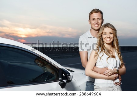 Lovely couple. Portrait of a beautiful blond girl with curvy hair and her handsome boyfriend hugging her from the back and smiling near their car, stunning sunset on the background