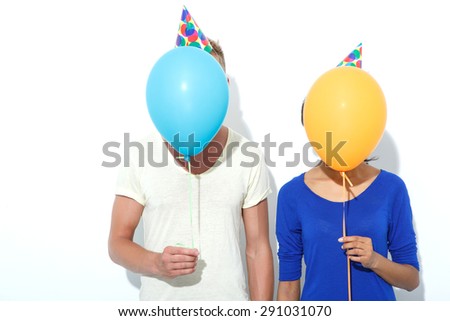 Mixed couple. Portrait of a blond young man and his beautiful mulatto girlfriend holding colorful balloons in front of their faces wearing colorful party hats, isolated on white background