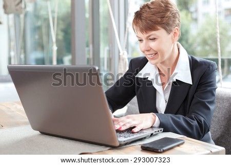 Portrait of a beautiful businesswoman wearing formal suit sitting at the table and working with a laptop her mobile phone lying near, in a restaurant during business lunch
