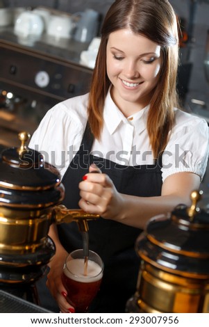 Portrait of a pretty bartender standing smiling and pouring beer, shelves full of bottles with alcohol on the background