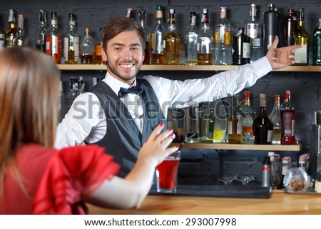 Portrait of a handsome bartender standing smiling and proposing some drinks to a young woman sitting back to us at the counter choosing, shelves full of bottles with alcohol on the background
