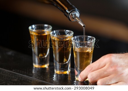 Close up photo of hands of a bartender pouring some drink into shot glasses on a wooden counter