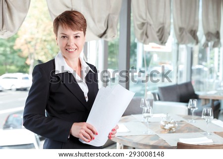 Waist up portrait of a beautiful businesswoman standing smiling and holding some documents in her hands, wearing a formal suit, in a restaurant during business lunch