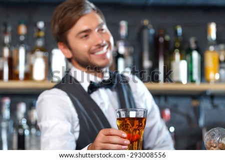 Portrait of a handsome bartender standing at the counter smiling and holding a glass of drink stretching it to us, shelves full of bottles with alcohol on the background