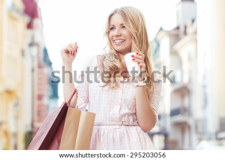 Portrait of a beautiful blond girl with long wavy hair wearing pink dress, standing on the street holding shopping bags  smiling and drinking coffee to go