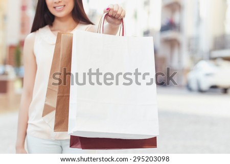 Portrait of a young girl wearing beige blouse and white jeans stretching her hand holding shopping bags in front of her, selective focus, beautiful old city on the background