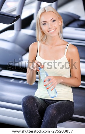 Portrait of a beautiful young blond girl wearing a white top and grey leggings sitting on the treadmill having some rest holding a bottle of still water, smiling in a fitness club