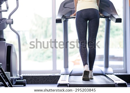 Half length photo of a fit young woman standing back to us on a treadmill, wearing grey leggings and white sneakers in a fitness club