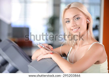 Close up portrait of a beautiful young blond girl wearing a white top looking at the camera and holding mp3 player listening to music, standing on a treadmill smiling in a fitness club
