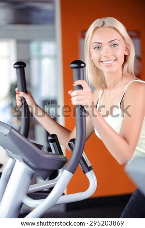 Portrait of a beautiful young blond girl wearing a white top looking at the camera, standing on elliptical trainer near a big window in a fitness club