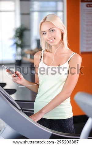 Portrait of a beautiful young blond girl wearing a white top looking at the camera and holding mp3 player listening to music, standing on a treadmill smiling in a fitness club