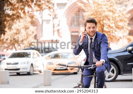Time to talk. Pleasant smiling bearded businessman looking aside and having nice conversation on mobile phone while riding bicycle.