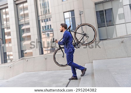 Time to ride. Vivacious businessman holding his bicycle on his shoulders and looking straight while going down stairs.