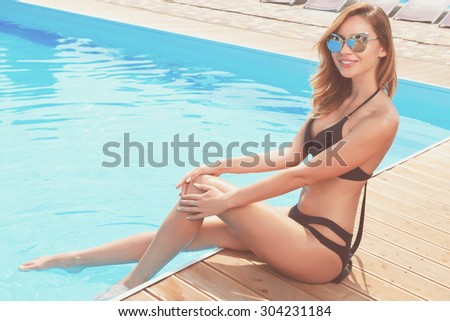 Feeling water. Portrait of young smiling red-haired woman wearing sunglasses sitting at poolside.