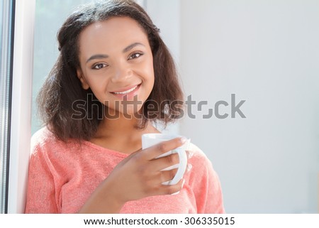 Love my tea. Happy young mulatto woman posing with cup of tea on background of window.