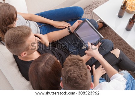 Look here. Top view of nice cheerful friends sitting on the sofa and holding laptop while watching news