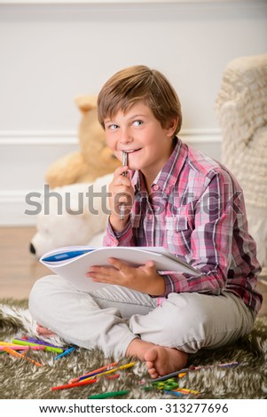 Let me think. Pleasant contented little boy sitting on the carpet and holding album with pen while thinking