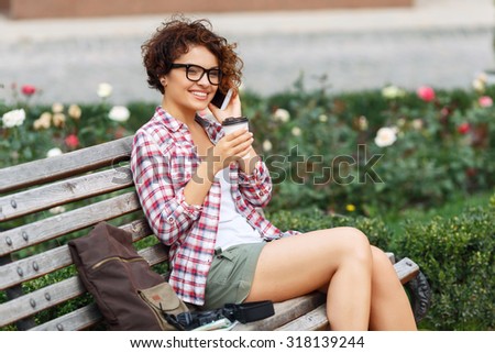 Live life fully. Pleasant blissful young girl talking on mobile phone and drinking coffee while sitting on the bench