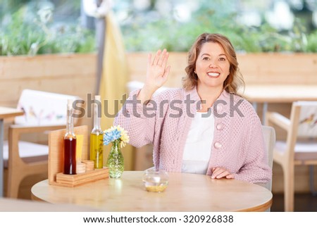 Nice to see you. Upbeat smiling charming adult woman holding her hand up and welcoming while sitting at the table in the cafe