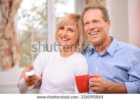 Time to rest. Overjoyed adult smiling couple bonding to each other and holding remote control while watching TV