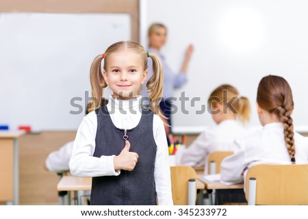 Young girl giving a thumbs up sign in a classroom with a teacher and other students in the background.