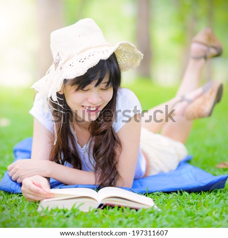 Beautiful happy Asian girl lies on meadow and reads the book , Happy smiling beautiful young university student studying lying down in grass.