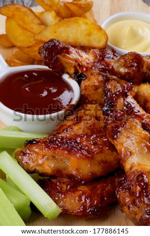 Juicy Chicken Legs and Wings Barbecue with French Fries, Ketchup, Cheese Sauce and Celery Sticks closeup on Wooden Board