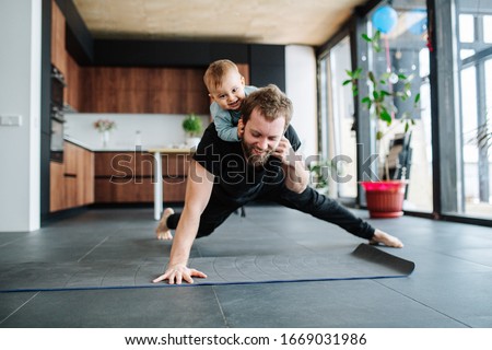 Father working out, doing single arm plank with his jolly infant baby riding on his neck. At home apartment.