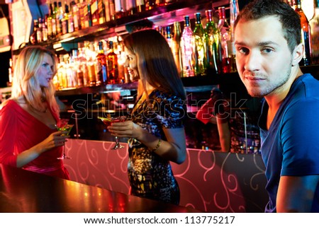 Portrait cute guy looking at camera at party with two girls chatting on background
