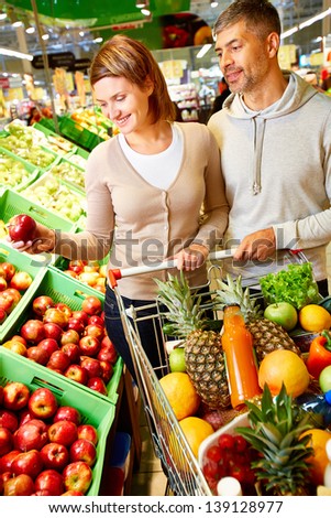 Image of happy couple with cart full of products choosing apples in supermarket