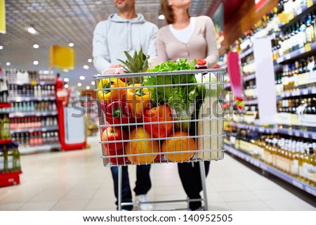 Image of cart full of products in supermarket being pushed by couple