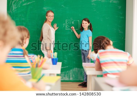 Portrait of smart teacher and schoolgirl standing by blackboard and looking at schoolkids in classroom