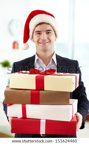 Image of happy businessman in Santa cap holding stack of gifts and looking at camera in office